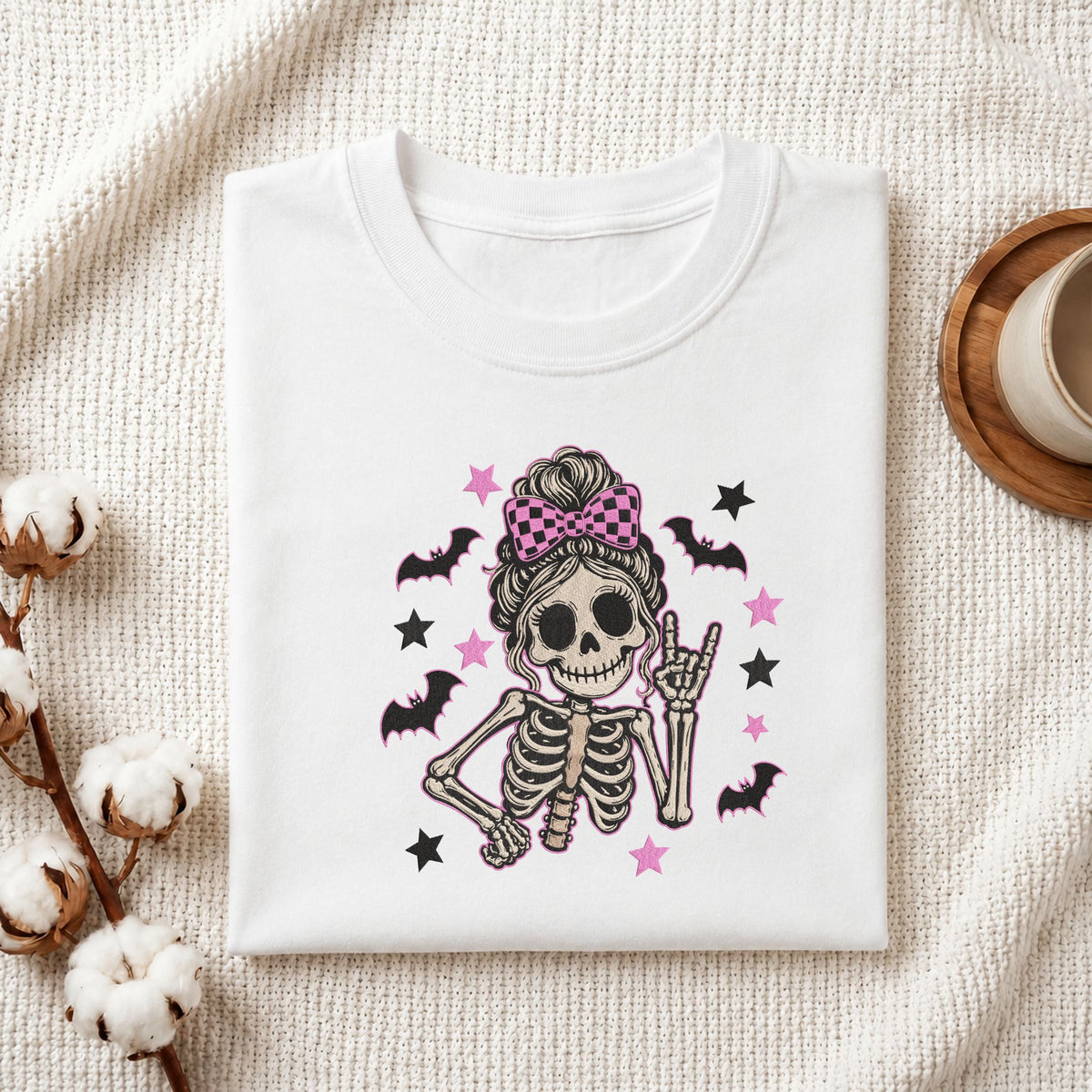 Folded white cotton t-shirt mockup featuring a cute skeleton messy bun machine embroidery design, styled with cotton stems and a wooden cup