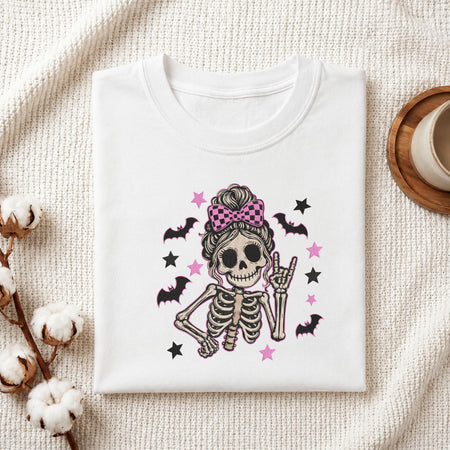 Folded white cotton t-shirt mockup featuring a cute skeleton messy bun machine embroidery design, styled with cotton stems and a wooden cup