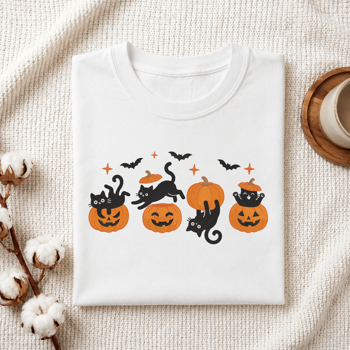 Folded white cotton t-shirt mockup featuring a playful black cats and pumpkins machine embroidery design, styled with cotton stems and a wooden cup