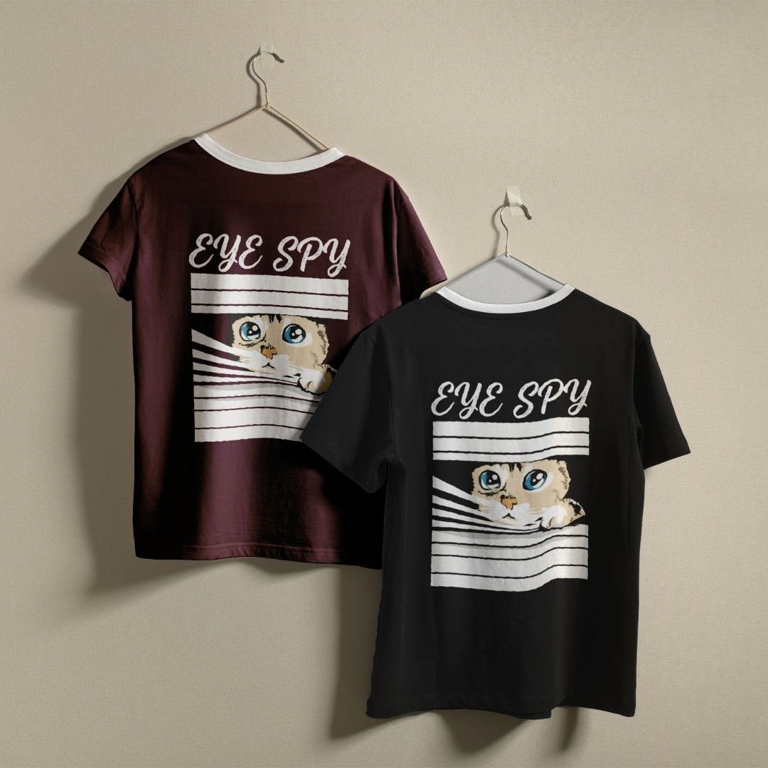 Two t-shirts hanging on a wall, one dark maroon and one black, both featuring the funny "EYE SPY" cat peeking through window blinds design.