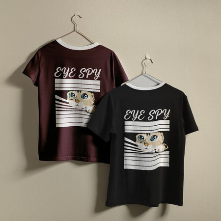 Two t-shirts hanging on a wall, one dark maroon and one black, both featuring the funny "EYE SPY" cat peeking through window blinds design.