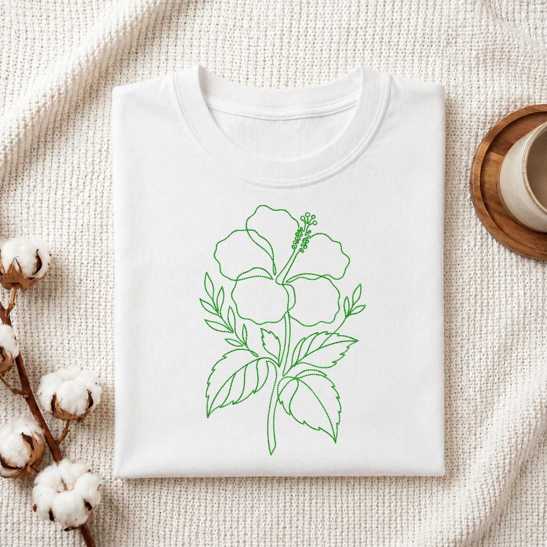 A folded white t-shirt featuring the green, line-art hibiscus embroidery design, presented on a textured white fabric background with cotton sprigs and a wooden coaster.