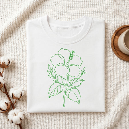 A folded white t-shirt featuring the green, line-art hibiscus embroidery design, presented on a textured white fabric background with cotton sprigs and a wooden coaster.