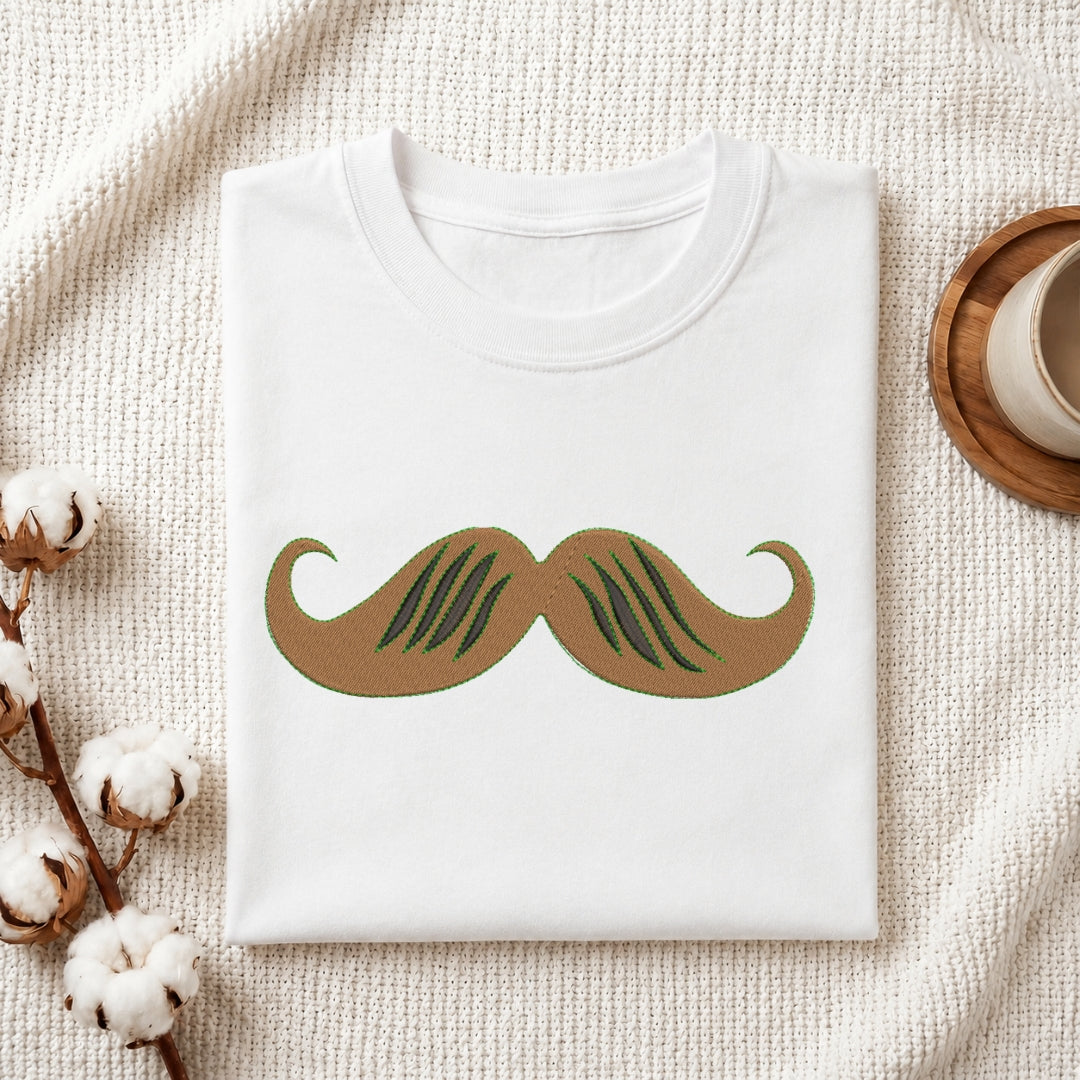 A folded white crewneck t-shirt with a stylized brown and green mustache graphic printed on the front, displayed on a textured cream knit blanket with cotton bolls and a wooden cup in the background.