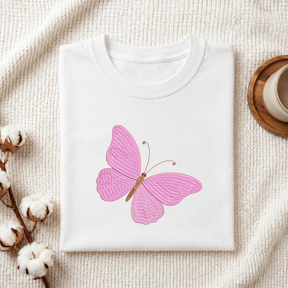 Folded white cotton t-shirt mockup featuring an elegant pink butterfly machine embroidery design, styled with cotton stems and a wooden cup.