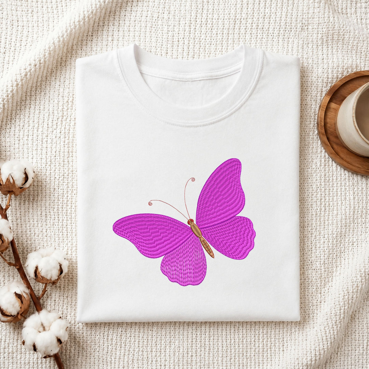 Folded white cotton t-shirt mockup featuring a vibrant purple butterfly machine embroidery design, styled with cotton stems and a wooden cup.