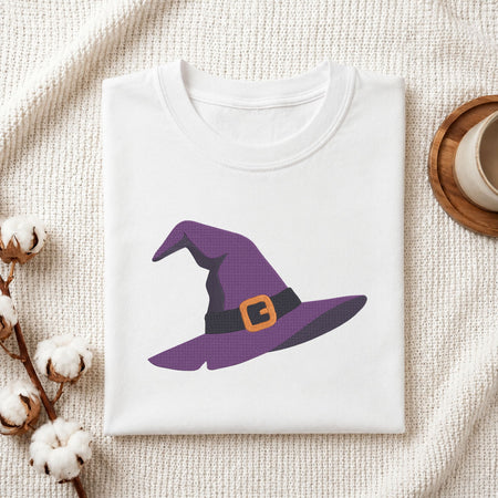 A folded white t-shirt with a purple witch hat graphic printed on the front, styled on a textured cream knit blanket with cotton bolls and a wooden cup in the background.