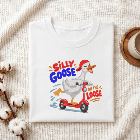 Folded white cotton t-shirt mockup featuring a "Silly Goose on the Loose" duck riding a red scooter machine embroidery design, styled with cotton stems and a wooden cup   