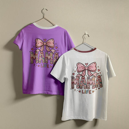 Two t-shirts hanging on a wall, one light purple and one white, both printed with a whimsical doodle-style "MAMA LIFE" floral letters and bow graphic.