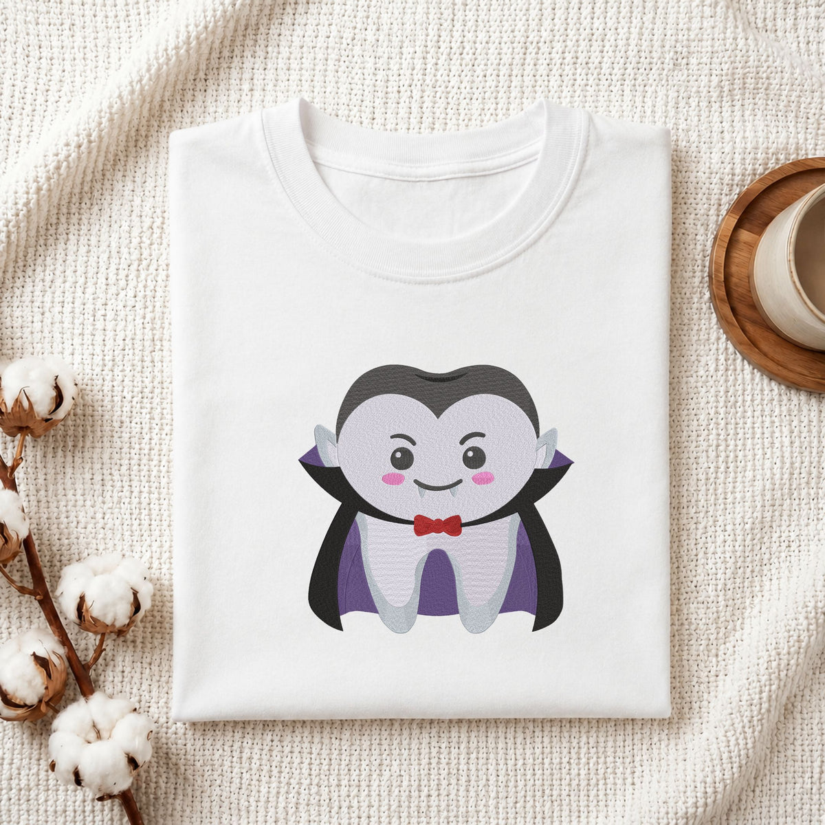 Folded white cotton t-shirt mockup featuring a cute vampire tooth Halloween dental machine embroidery design, styled with cotton stems and a wooden cup