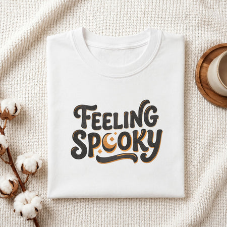 Folded white cotton t-shirt mockup featuring a "Feeling Spooky" machine embroidery design, styled with cotton stems and a wooden cup.