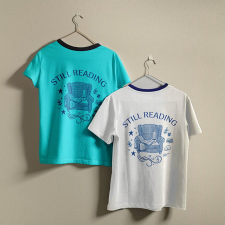 Two t-shirts hanging on a wall, one turquoise blue and one white, both featuring the retro line art "Still Reading" cozy armchair graphic design.
