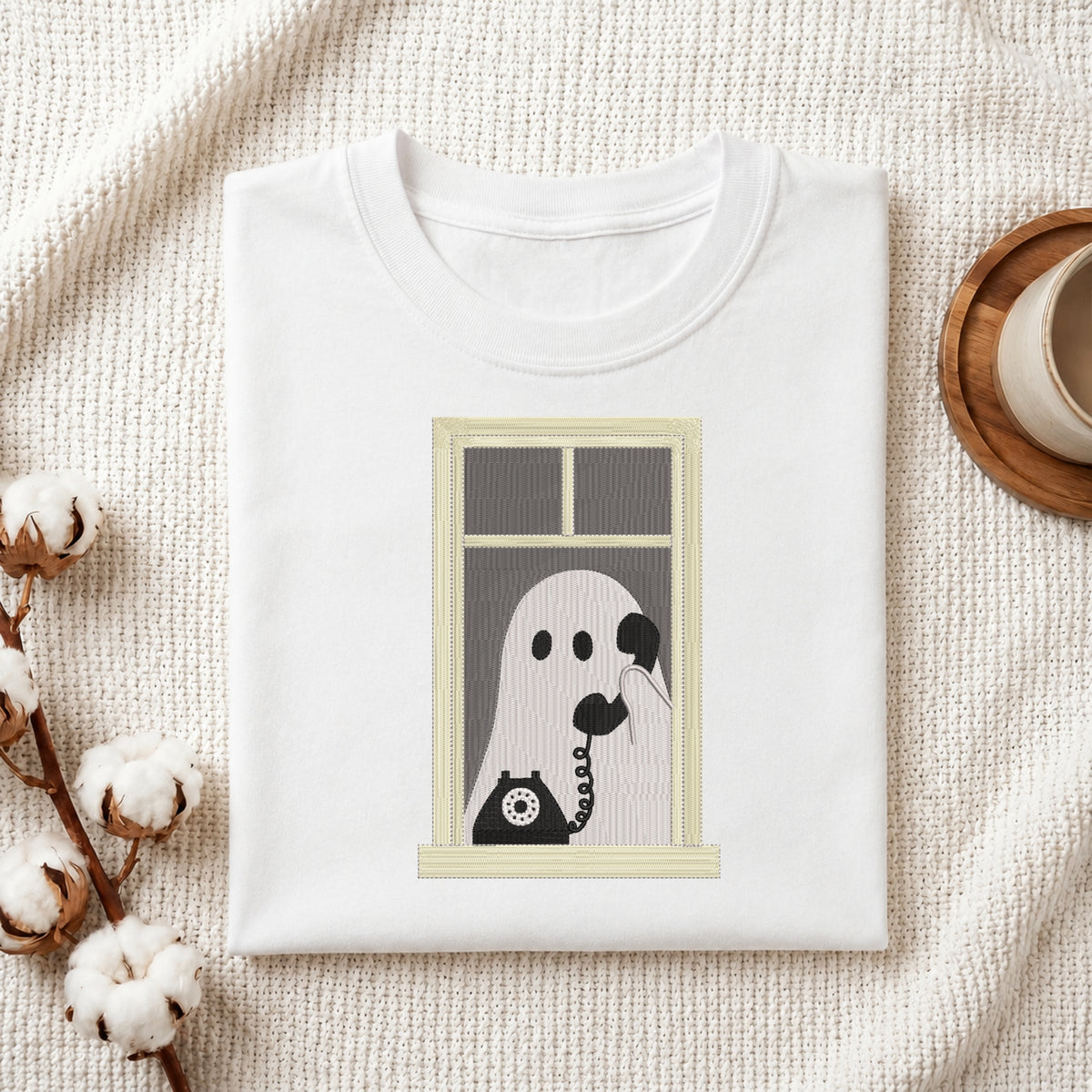 A folded white t-shirt with a graphic print of a ghost on a telephone hanging from a window, styled on a textured cream fabric with cotton bolls and a wooden cup.