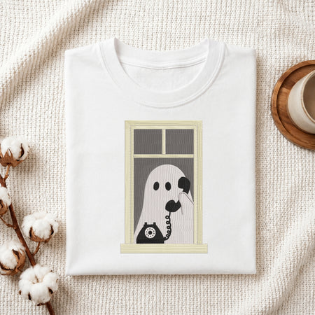 A folded white t-shirt with a graphic print of a ghost on a telephone hanging from a window, styled on a textured cream fabric with cotton bolls and a wooden cup.