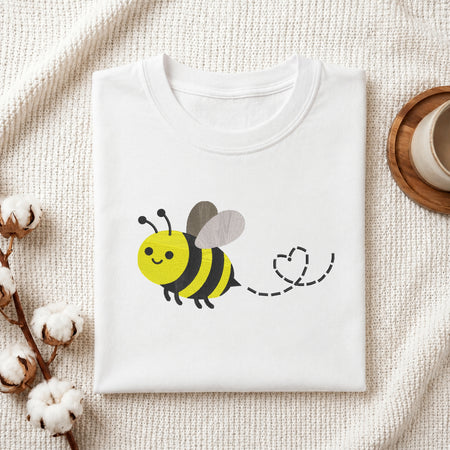 A folded white t-shirt with a cute cartoon bee graphic on the front, styled on a textured cream knit blanket with cotton bolls and a wooden cup in the background.