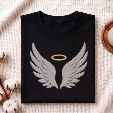 A folded black crewneck t-shirt featuring a large embroidered design of gray angel wings with a gold halo, displayed on a textured beige fabric background with decorative cotton bolls.