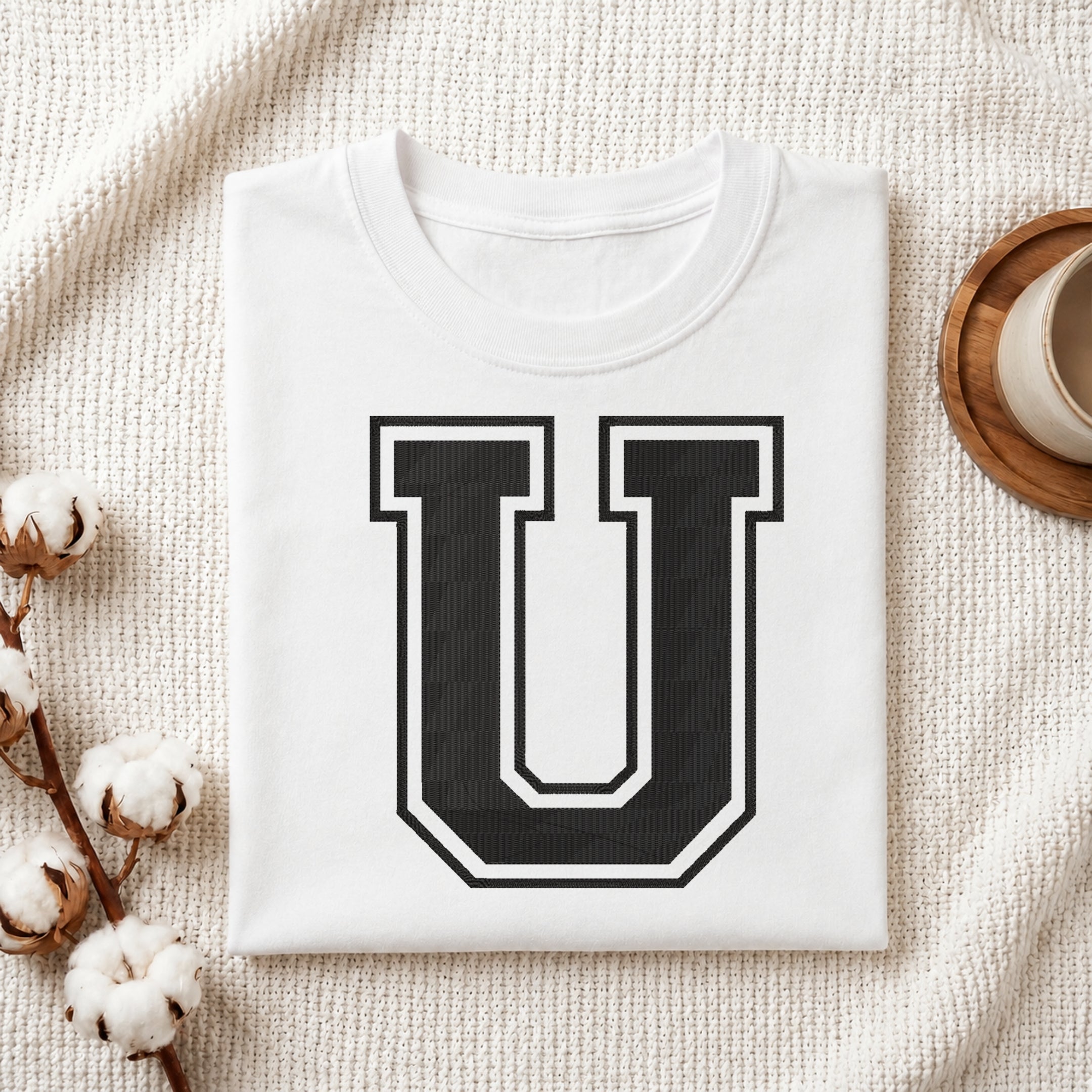 A folded white t-shirt with a large black and white block letter 'U' printed on the front, styled in a flat lay on a textured cream fabric with cotton bolls and a cup on a wooden coaster.