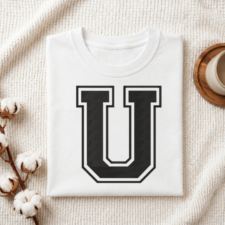 A folded white t-shirt with a large black and white block letter 'U' printed on the front, styled in a flat lay on a textured cream fabric with cotton bolls and a cup on a wooden coaster.