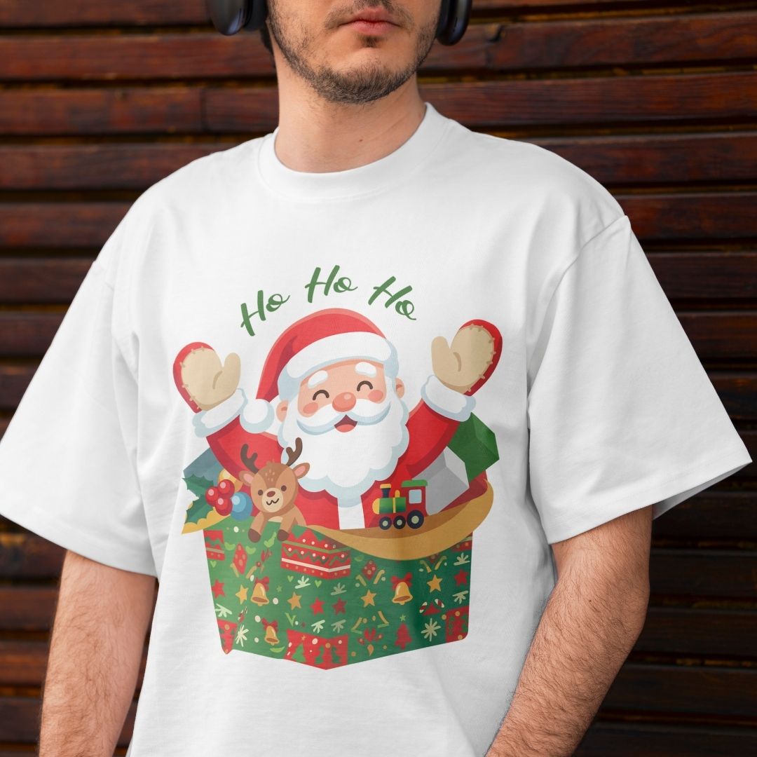 A man wearing a white crewneck t-shirt with a colorful graphic of Santa Claus popping out of a gift box with the text 'Ho Ho Ho' above him, set against a dark wooden plank background.