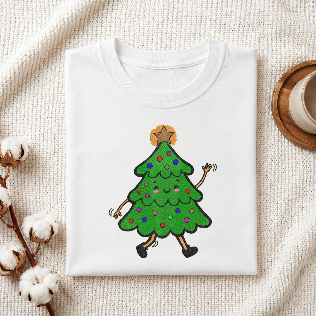 A folded white crewneck t-shirt featuring a cartoon graphic of a walking Christmas tree with colorful ornaments and a gold star on its head, displayed on a textured cream fabric background with decorative cotton bolls and a coffee cup.