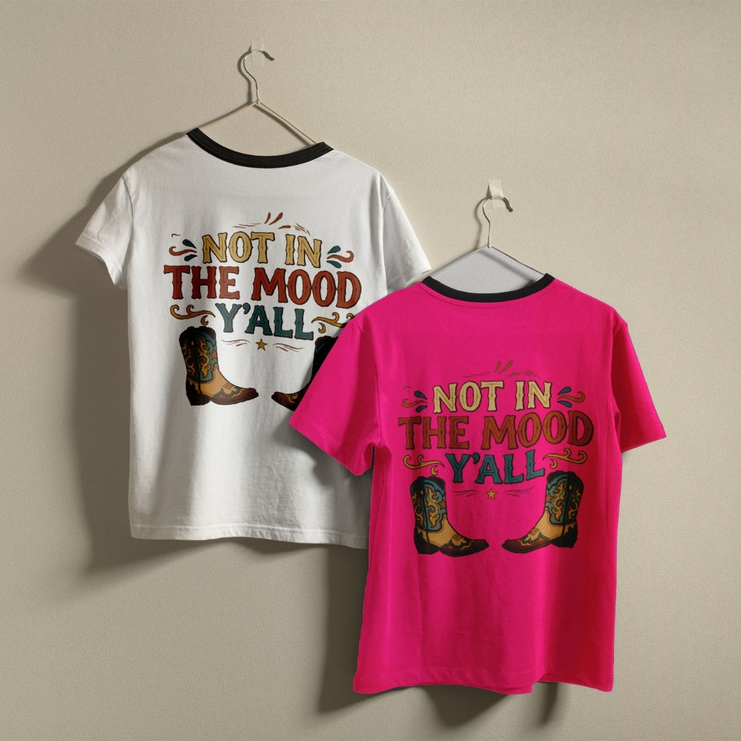 Two ringer t-shirts with black neck trims hanging on metal hangers against a beige wall. The left shirt is white with a western-style graphic reading 'NOT IN THE MOOD Y'ALL' in brown and teal, with cowboy boots.