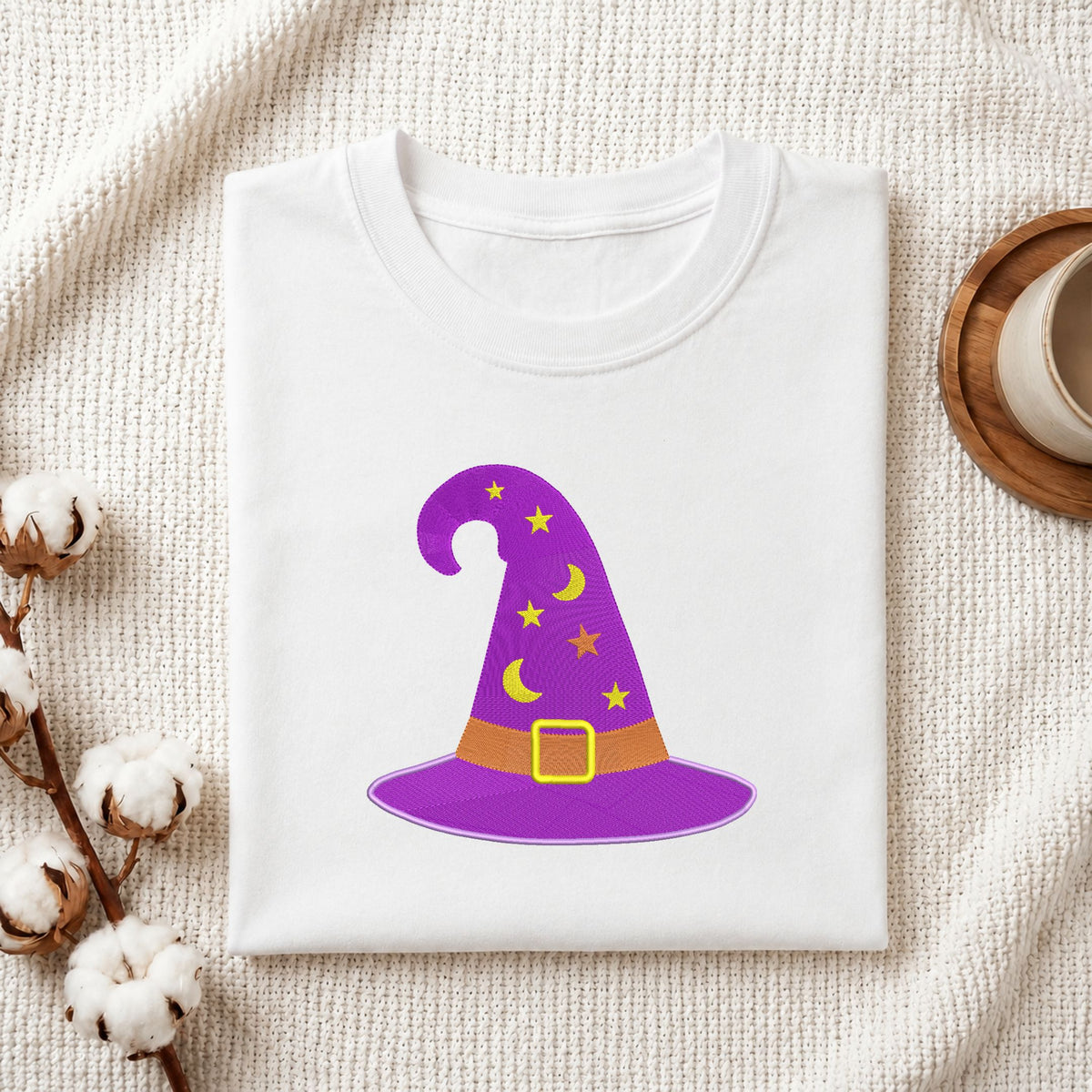 Folded white cotton t-shirt mockup featuring a purple witch hat machine embroidery design, styled with cotton stems and a wooden cup.
