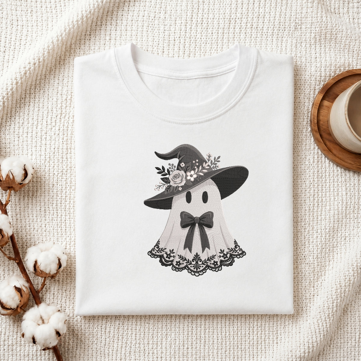 A folded white crewneck t-shirt featuring a cute black and white graphic of a ghost wearing a witch hat with flowers, styled on a textured cream fabric with cotton bolls and a wooden cup.