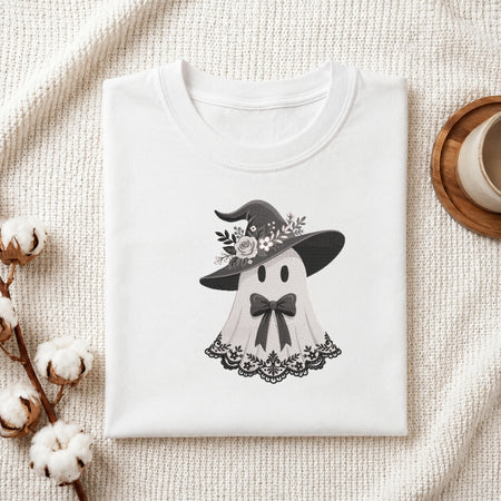 A folded white crewneck t-shirt featuring a cute black and white graphic of a ghost wearing a witch hat with flowers, styled on a textured cream fabric with cotton bolls and a wooden cup.