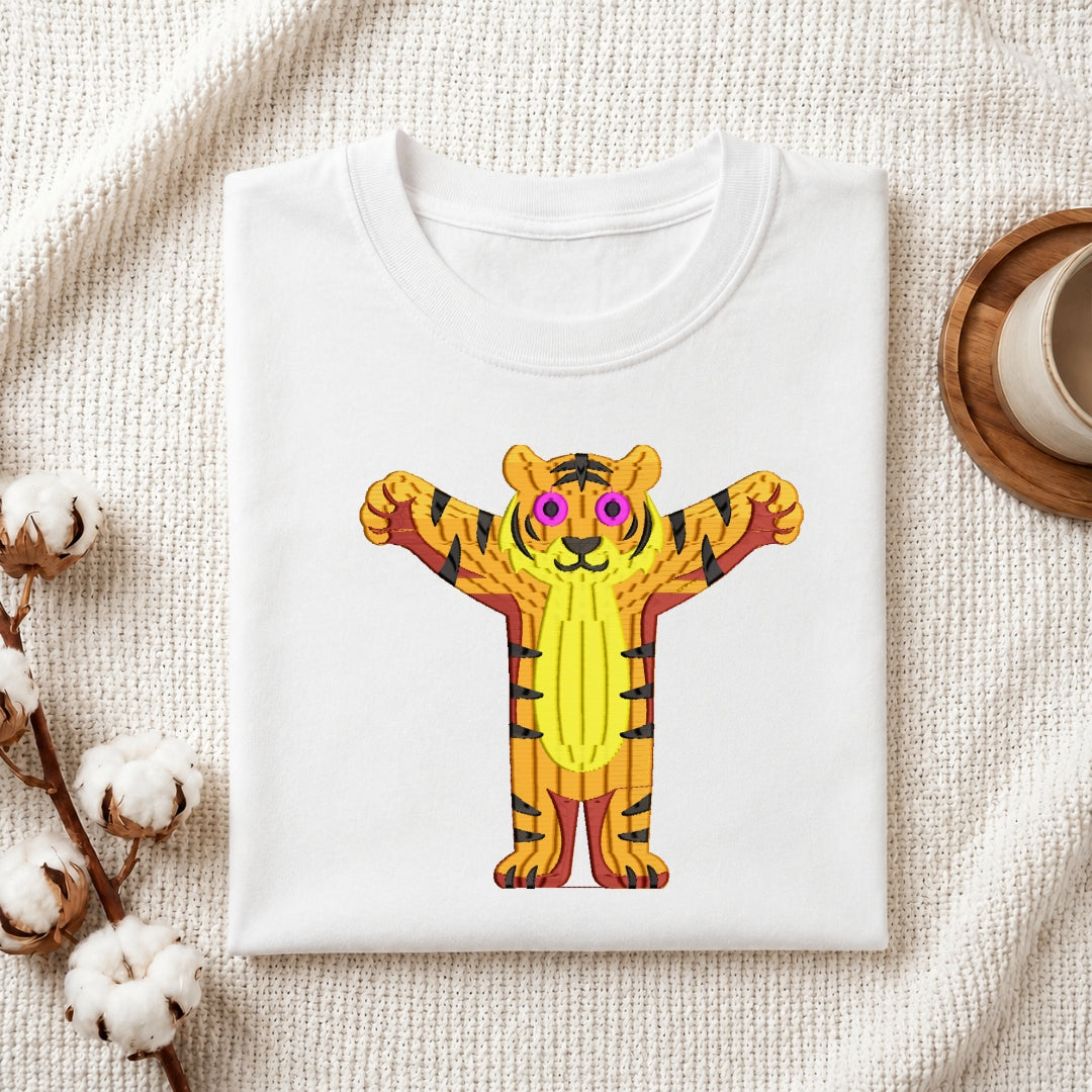 A folded white crewneck t-shirt featuring a colorful graphic of a cartoon tiger with pink sunglasses and raised paws, styled on a textured cream fabric with cotton bolls and a wooden cup.