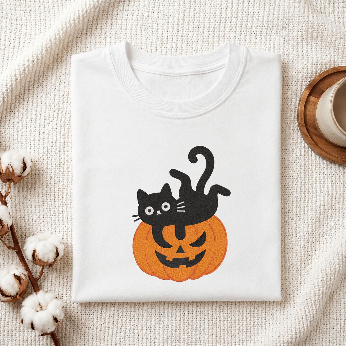 Folded white cotton t-shirt mockup featuring a cute black cat lying on a jack-o-lantern pumpkin machine embroidery design, styled with cotton stems and a wooden cup