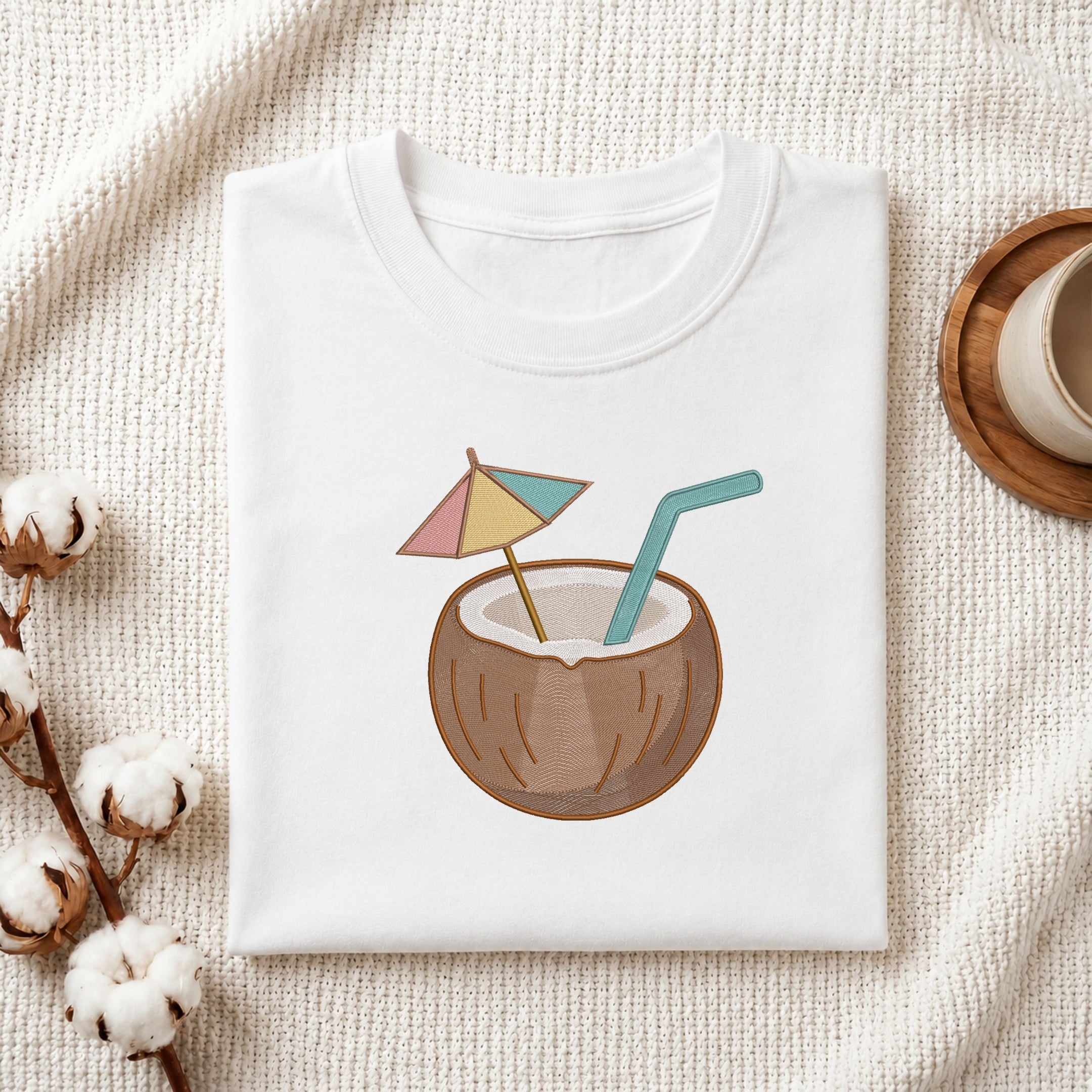 A folded white t-shirt with a cartoon coconut drink graphic, styled on a textured cream fabric with cotton bolls and a wooden cup.