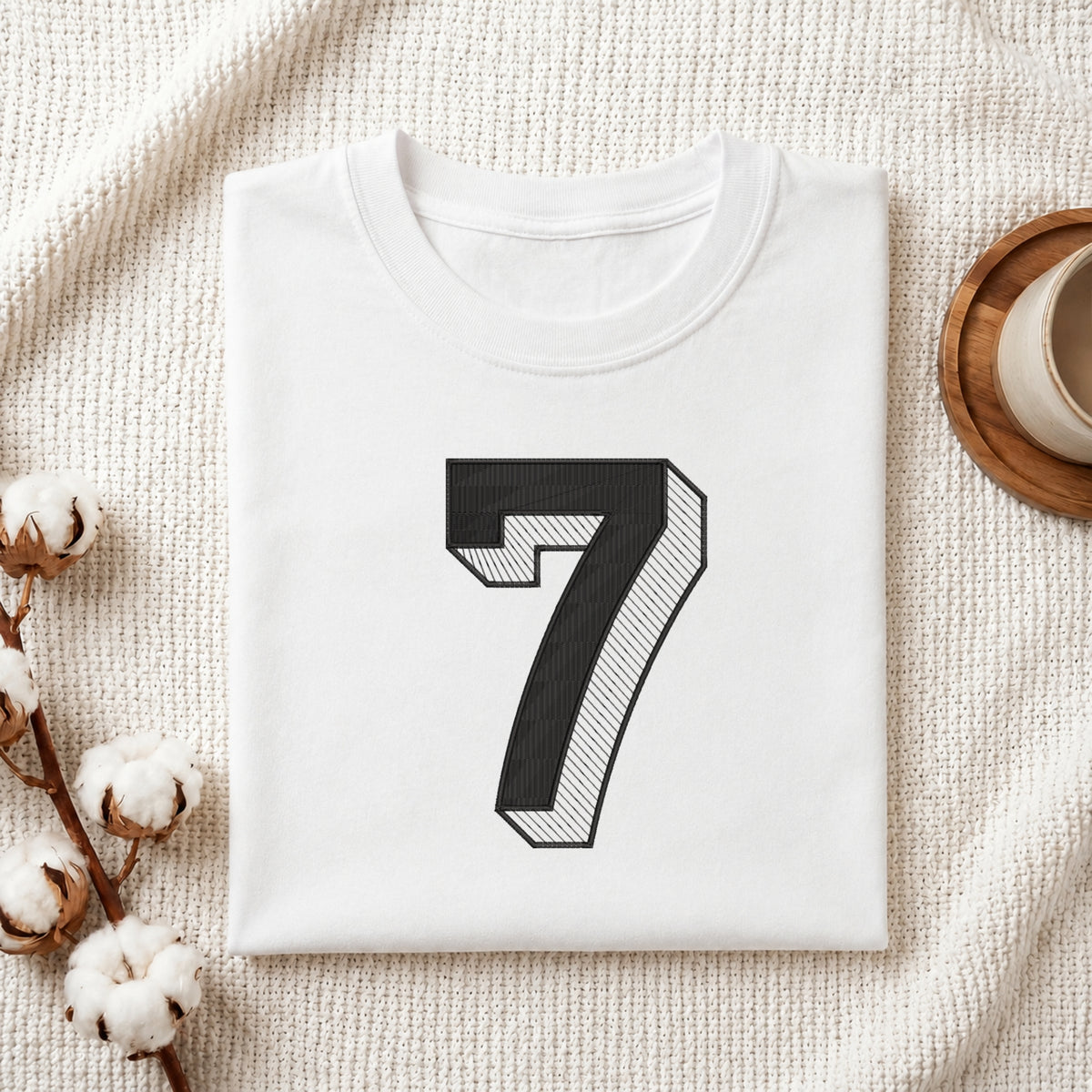 A folded white crewneck t-shirt with a large black number 7 printed on the front, styled on a textured cream blanket with cotton bolls and a cup in the background.