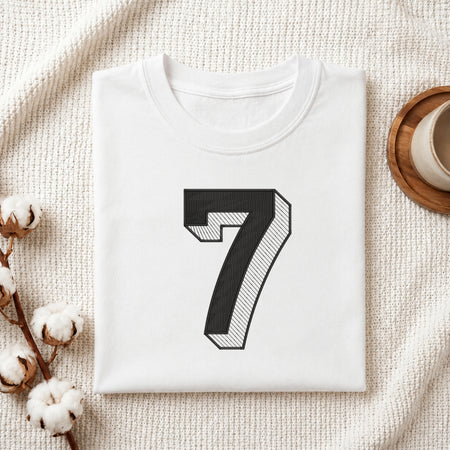 A folded white crewneck t-shirt with a large black number 7 printed on the front, styled on a textured cream blanket with cotton bolls and a cup in the background.