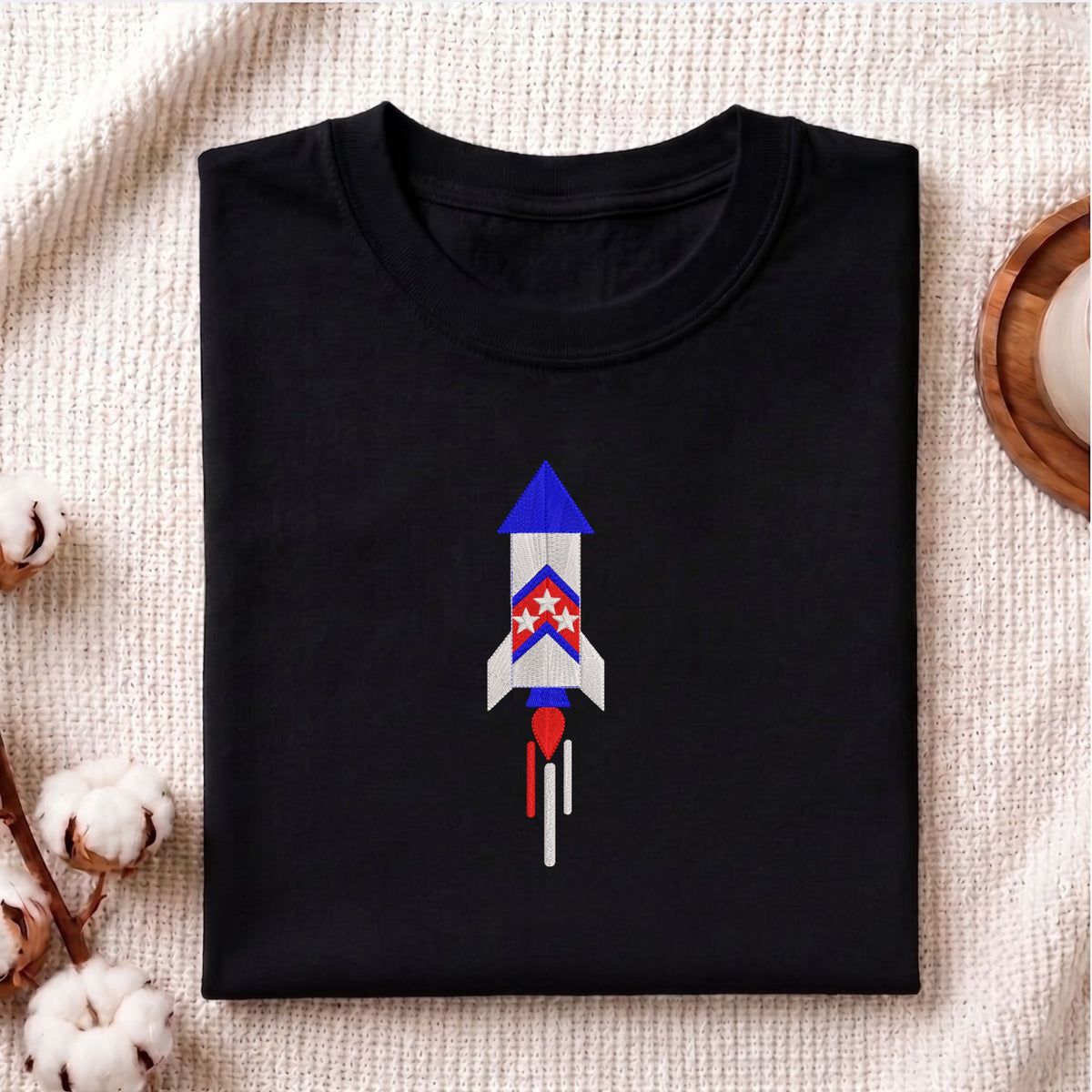 Folded black cotton t-shirt mockup featuring a patriotic space rocket machine embroidery design, styled with cotton stems and a wooden cup.   