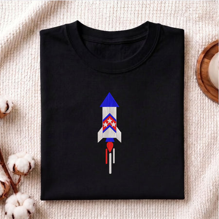 Folded black cotton t-shirt mockup featuring a patriotic space rocket machine embroidery design, styled with cotton stems and a wooden cup.   