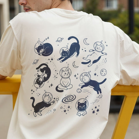 A person wearing a cream-colored t-shirt, viewed from the back. The shirt features a large graphic print of various cartoon cats in space outfits, including helmets and spacesuits, with stars, planets, and moons in dark blue.