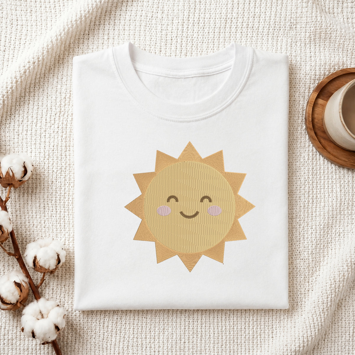 A folded white crewneck t-shirt featuring a cute embroidered-style smiling sun graphic in shades of yellow and beige with pink cheeks, styled on a textured cream knit blanket with cotton bolls and a wooden cup in the background.