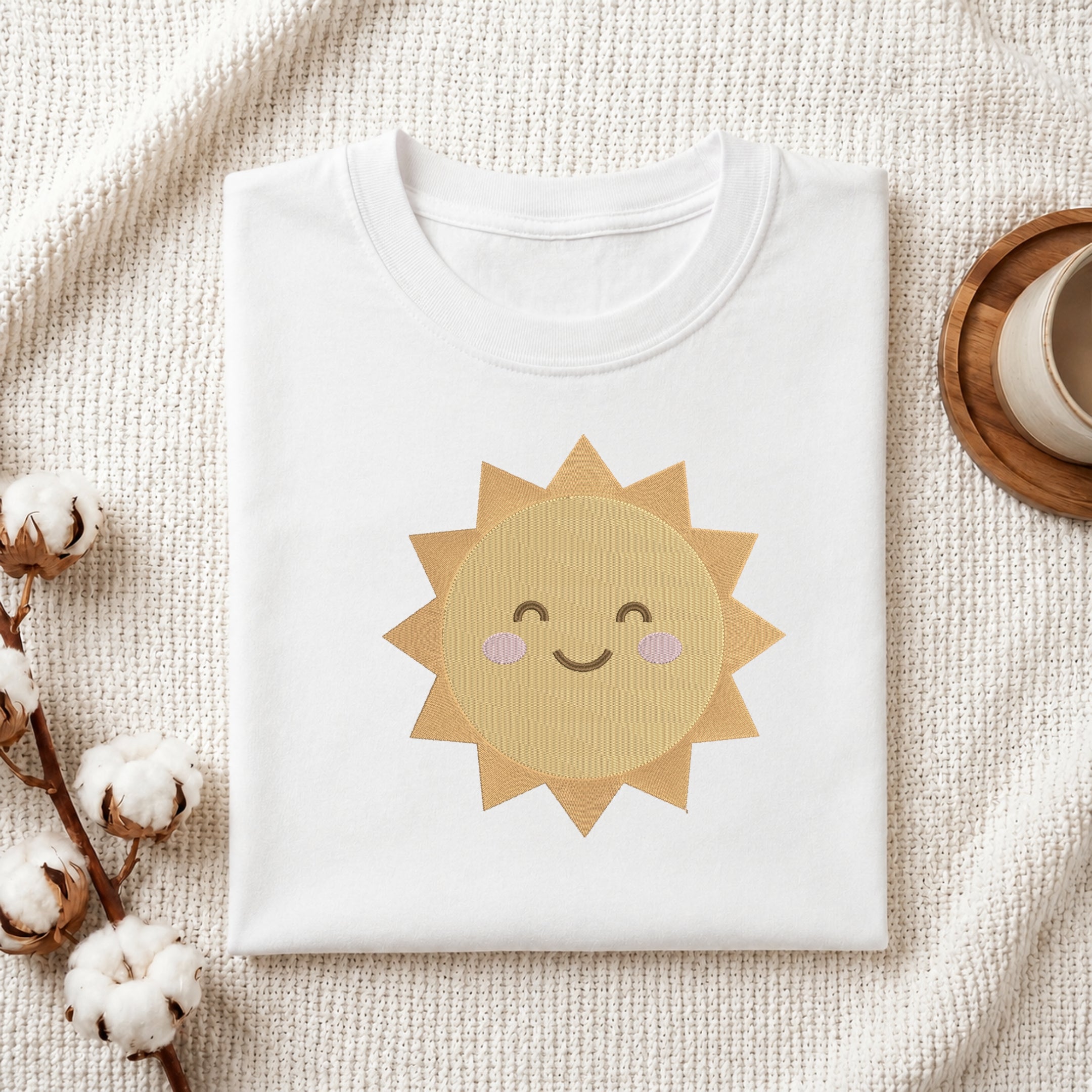 A folded white crewneck t-shirt featuring a cute embroidered-style smiling sun graphic in shades of yellow and beige with pink cheeks, styled on a textured cream knit blanket with cotton bolls and a wooden cup in the background.