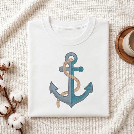 A folded white crewneck t-shirt featuring a graphic of a blue anchor with a beige rope wrapped around it, styled on a textured cream fabric with cotton bolls and a wooden cup.
