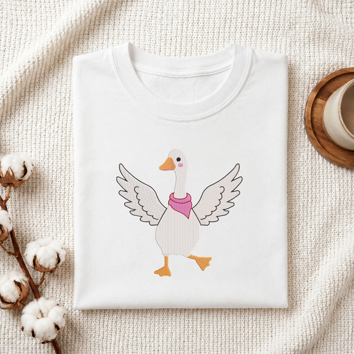 Folded white cotton t-shirt mockup featuring a cute dancing goose machine embroidery design, styled with cotton stems and a wooden cup