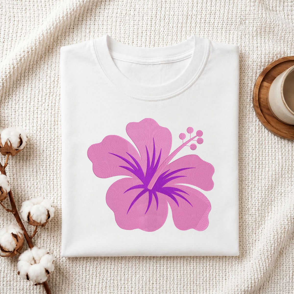 A folded white crewneck t-shirt featuring a large pink and purple hibiscus flower graphic on the front, styled on a textured cream knit blanket with cotton bolls and a wooden cup in the background.