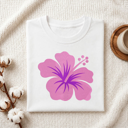 A folded white crewneck t-shirt featuring a large pink and purple hibiscus flower graphic on the front, styled on a textured cream knit blanket with cotton bolls and a wooden cup in the background.