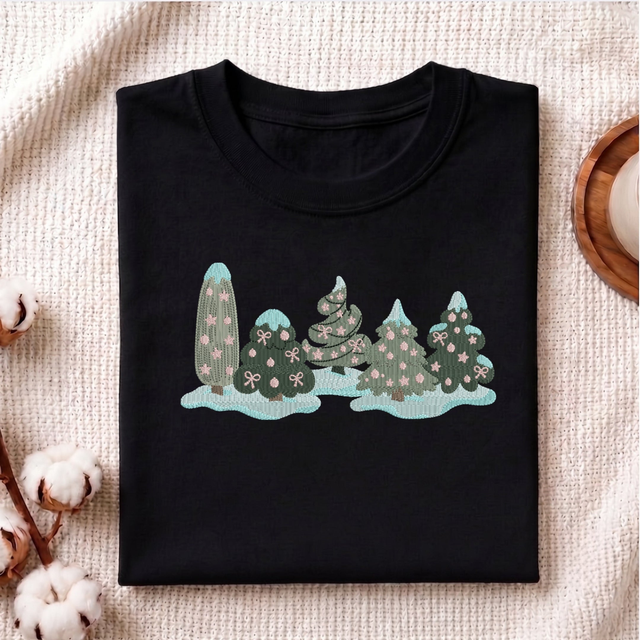 A folded black crewneck t-shirt with a graphic of stylized pine trees in shades of teal, green, and pink, displayed on a textured beige fabric background with decorative cotton bolls.