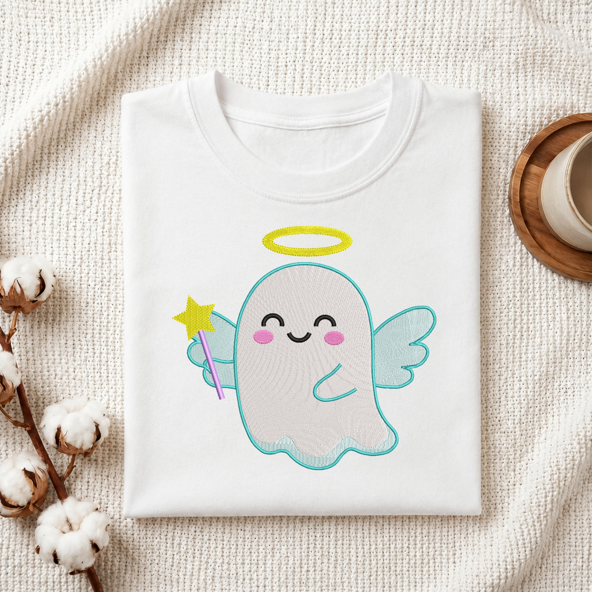A folded white crewneck t-shirt featuring a cute cartoon ghost graphic with a yellow halo, pink cheeks, and a purple wand, styled on a textured cream fabric with cotton bolls and a coffee cup.