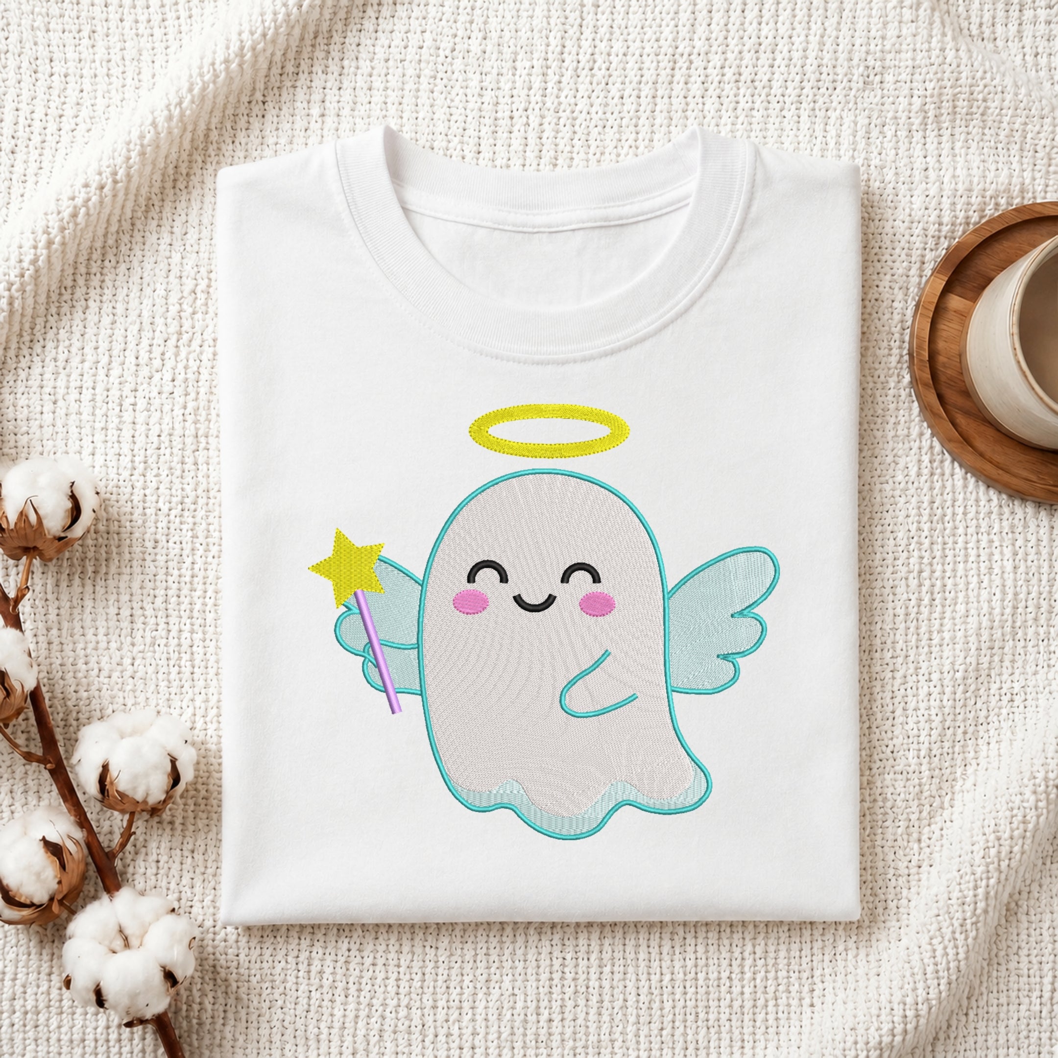 A folded white crewneck t-shirt featuring a cute cartoon ghost graphic with a yellow halo, pink cheeks, and a purple wand, styled on a textured cream fabric with cotton bolls and a coffee cup.