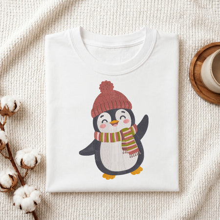 A folded white crewneck t-shirt featuring a cute cartoon penguin wearing a red knit hat and a red, green, and white striped scarf. The t-shirt is styled in a cozy flat-lay setting on a textured cream knit blanket, with cotton bolls on the left and a wooden cup on the right.
