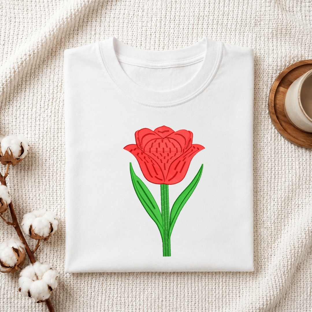 A folded white crewneck t-shirt featuring a graphic of a red tulip with green leaves, styled on a textured cream fabric with cotton bolls and a wooden cup in the background.