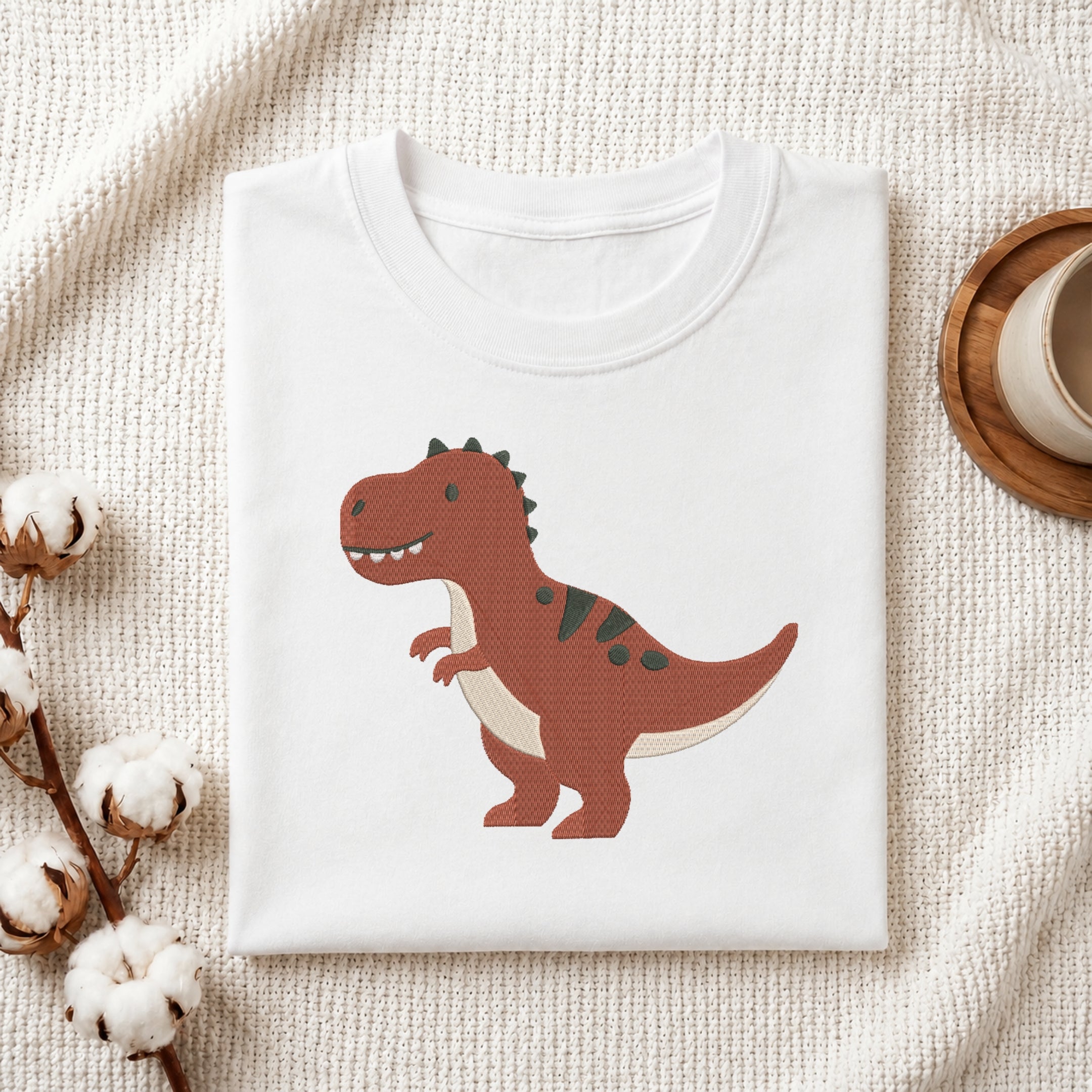 A folded white crewneck T-shirt displayed on a textured cream fabric background, featuring a cartoon-style brown T-rex dinosaur graphic on the front. The scene is styled with cotton bolls on the left and a wooden cup on the right.
