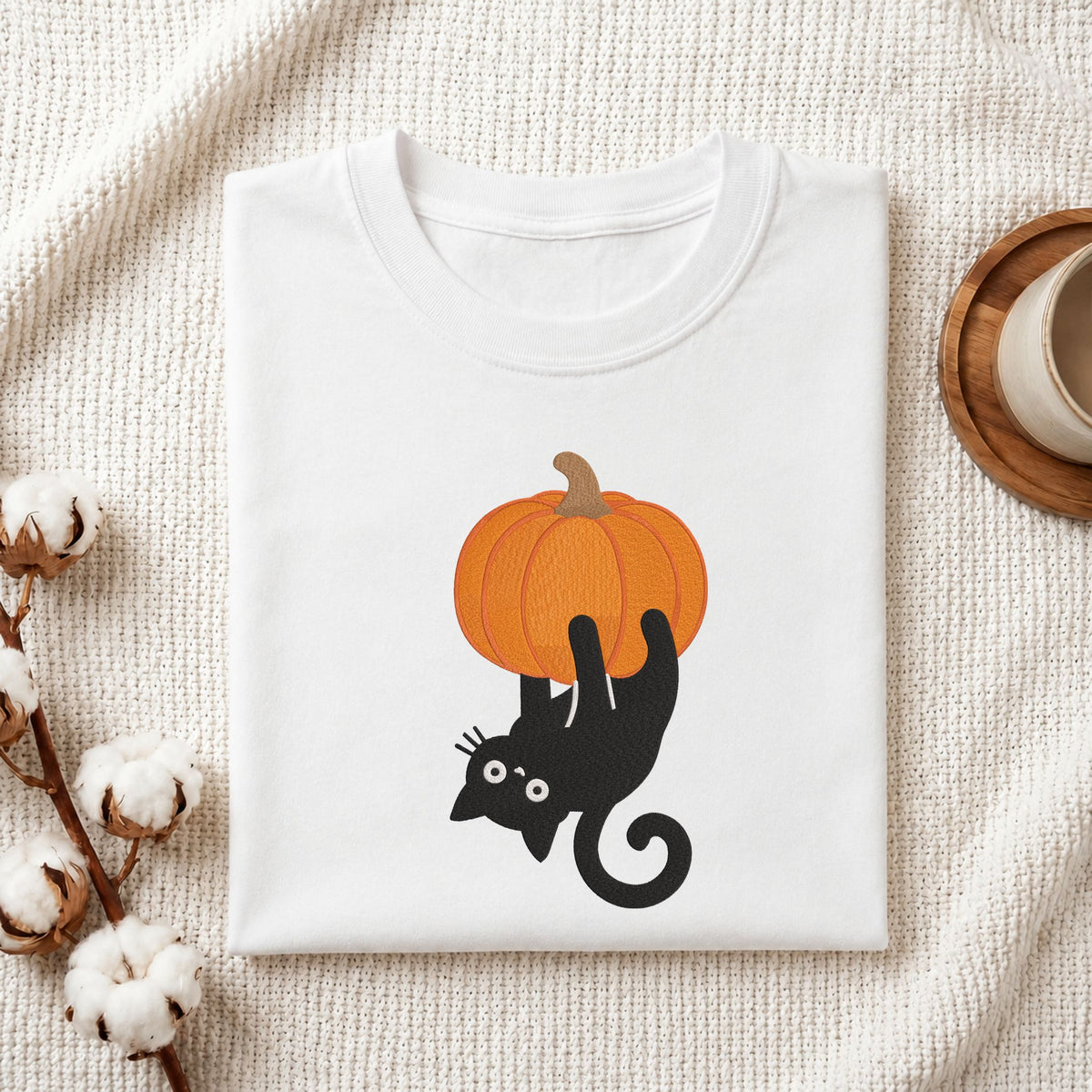 Folded white cotton t-shirt mockup featuring a mischievous black cat hanging upside down from a pumpkin machine embroidery design, styled with cotton stems and a wooden cup