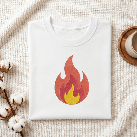 A folded white crewneck t-shirt with a stylized flame graphic in red, orange, and yellow on the front, presented on a textured cream knit blanket with cotton bolls and a wooden cup in the background.